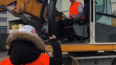 Foto von zwei Personen, die eine sitzt in einem Bagger, die andere steht davor