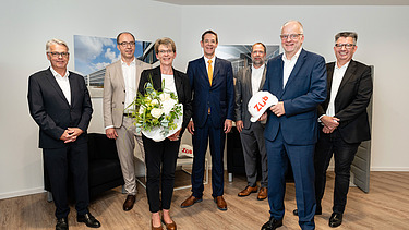 Foto von mehren Personen die schick gekleidet sind. Sie schauen freundlich. Vorne steht eine Frau mit Blumen in der Hand. 