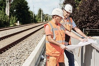 Foto von einem Bahngleis, rechts stehen zwei Personen in Schutzkleidung, die einen Plan in der Hand halten
