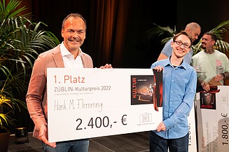 Foto von zwei Personen, die freundlich schauen. Sie halten den Preis zum ersten Platz des Comedy-Battles. 