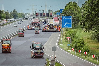 Foto einer Autobahnbaustelle mit mehreren Straßenbaumaschinen darauf, rechts ein blaues Verkehrsschild