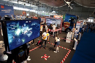 Foto von einem Großbildschirm in einer Messehalle, davor stehen zwei Personen