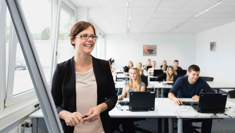 Foto eines Raumes mit mehreren sitzenden Personen, im Vordergrund steht eine weibliche Person mit Brille