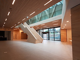 Foto vom inneren eines Gebäudes in der Eingangshalle. In der Mitte erstreckt sich eine Treppe aus Holz.  