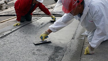  Foto von zwei Personen die mit Spachteln ein Gitter mit Beton bestreichen.