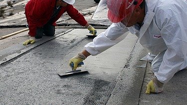  Foto von zwei Personen die mit Spachteln ein Gitter mit Beton bestreichen.