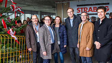 Foto von freundlich schauenden Personen auf einer Baustelle die neben einem Tannenbaum stehen.  