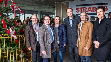 Foto von freundlich schauenden Personen auf einer Baustelle die neben einem Tannenbaum stehen.  