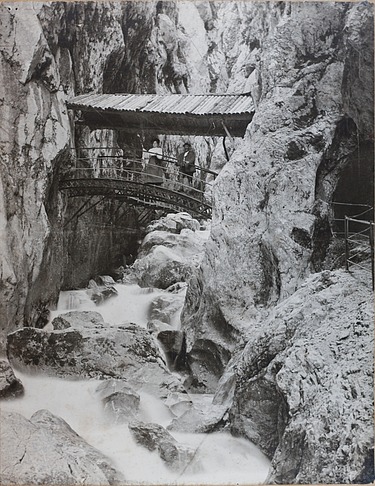 Schwarz-weiß Foto von einer Brücke, die über einen Graben geht. Drumherum sind Bergwände, die sich zu einer Schlucht formen. Auf der Brücke sind zwei Personen zu sehen. 