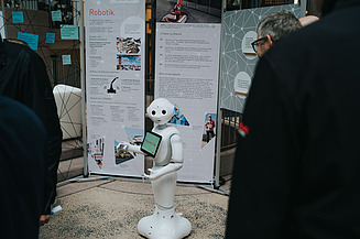Photo of a small, white robot standing in front of a trade fair booth
