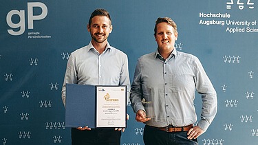 Foto von zwei Personen vor blauem Hintergrund, die linke Person hält eine Urkunde