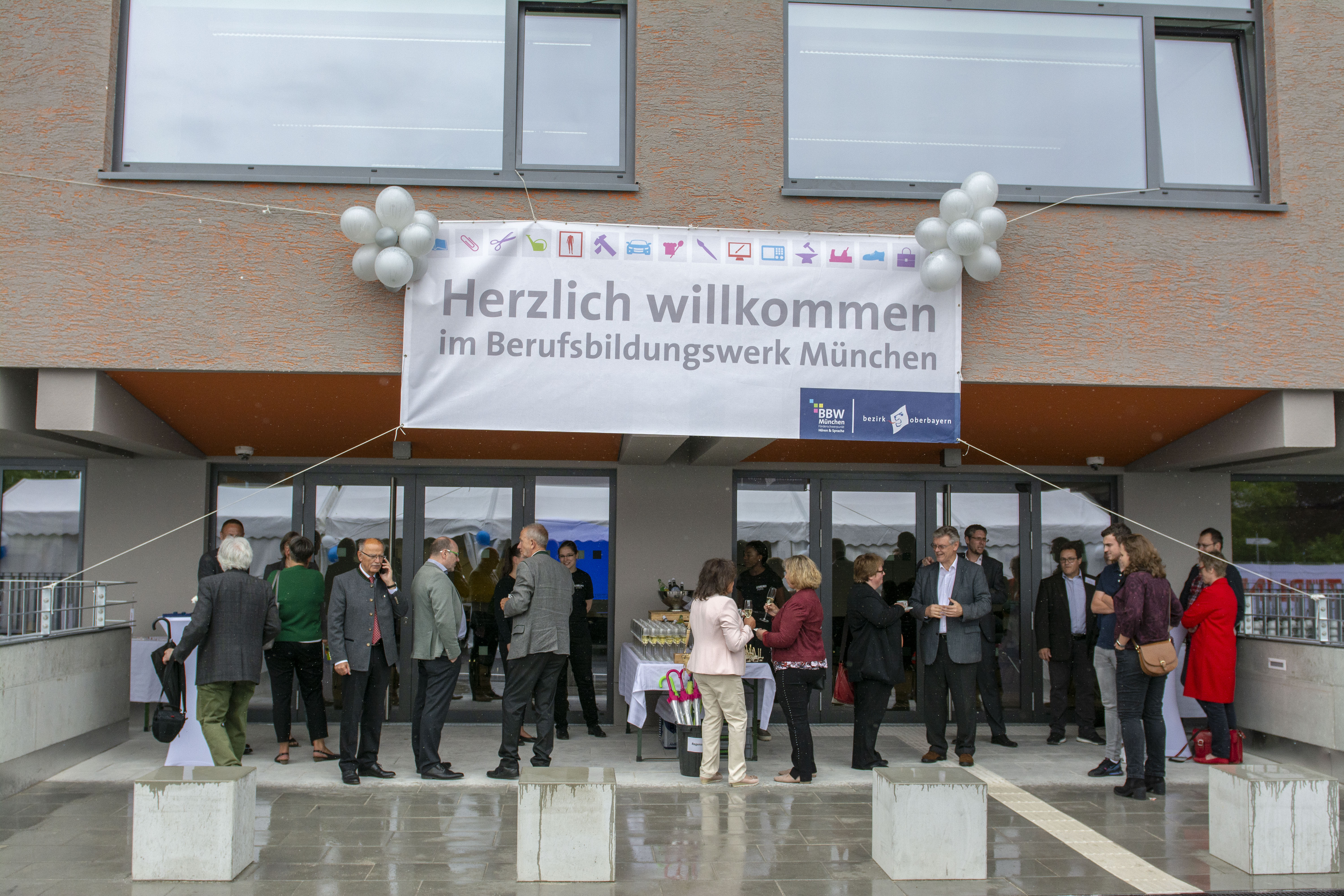 Foto vom Eingang des Berufsbildungswerk München.  Es vorm Eingang. Es herrscht eine ausgelassene Stimmung. 