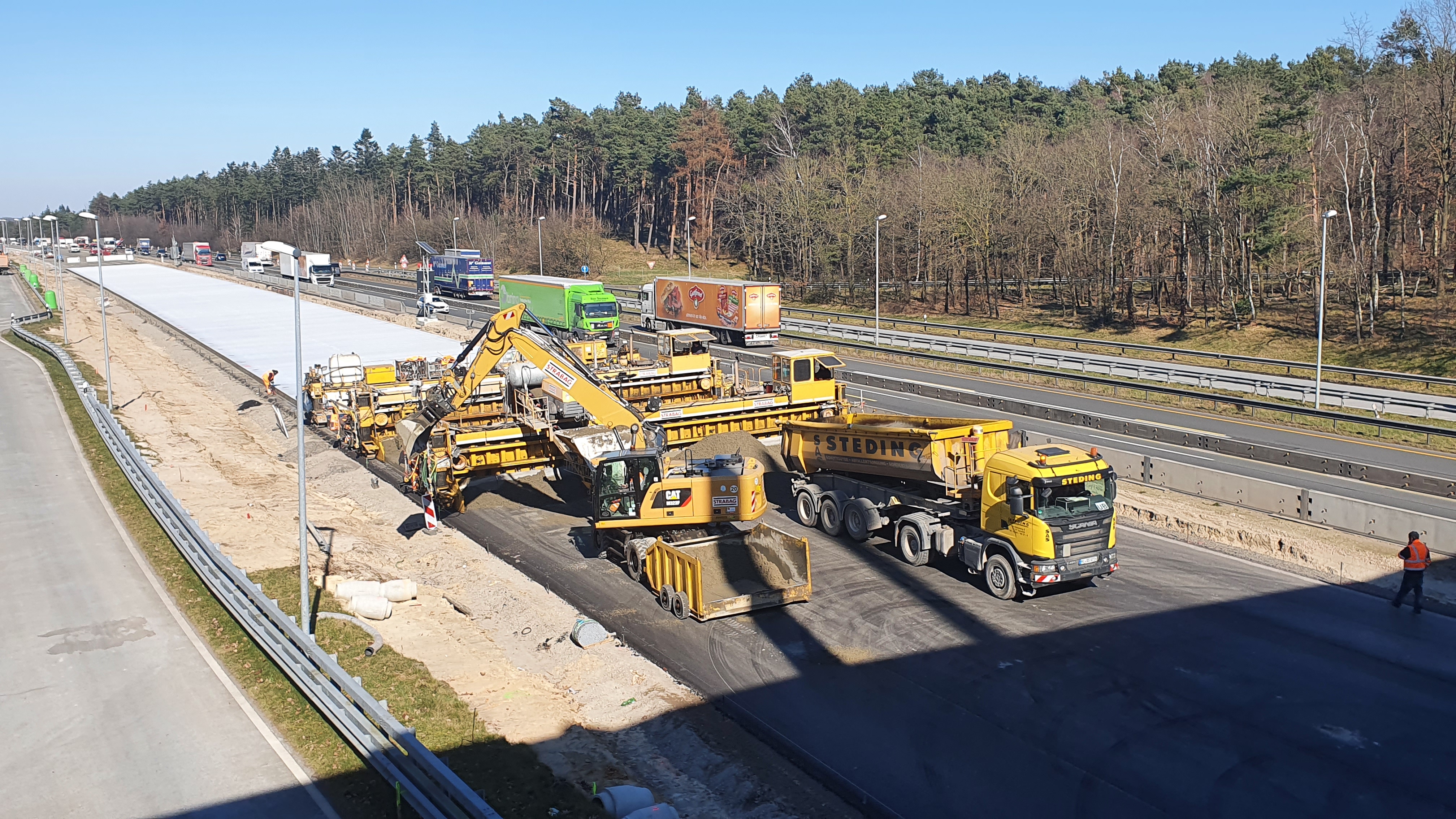 Das Bild zeigt eine nähere Ansicht von mehreren Straßenbaumaschinen beim Einbau einer Betondecke