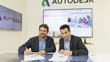 Foto von zwei Personen bei einer Vertragsunterzeichung, dahinter ein Bildschirm, darüber die Aufschrift Autodesk