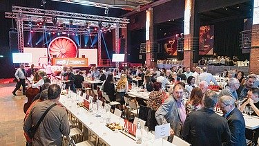 Foto von einer großen Anzahl an feiernden Personen in einem Festsaal, sitzend an weißgedeckten Tischen, im Hintergrund eine Bühne mit Scheinwerfern