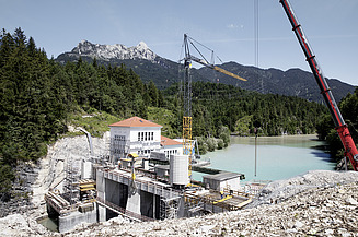 The photo shows the construction site of a dam in Tyrol in front of a lake in a wooded area.