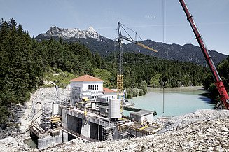 The photo shows the construction site of a dam in Tyrol in front of a lake in a wooded area.