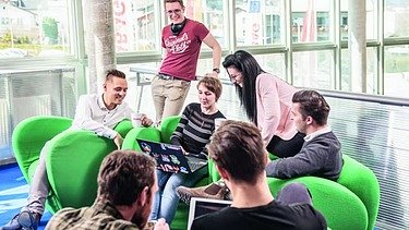 Das Bild zeigt eine Gruppe von jungen Personen auf grünen Stoffsesseln, im Hintergrund Glaswände