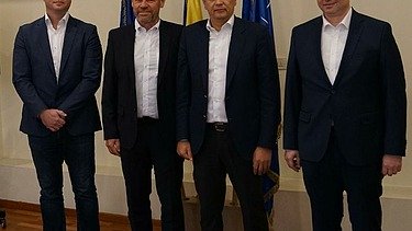 Foto von vier stehenden Personen vor drei Flaggen