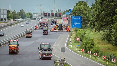 Foto einer Autobahnbaustelle mit mehreren Straßenbaumaschinen darauf, rechts ein blaues Verkehrsschild