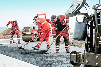 Foto von drei Straßenbauarbeitern mit Besen bei der Arbeit, rechts im Vordergrund sieht man einen Teil einer Straßenbaumaschine