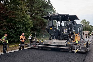 Foto von einer Asphalteinbaumaschine beim Einsatz, einige Personen stehen an beiden Seiten
