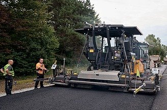 Foto von einer Asphalteinbaumaschine beim Einsatz, einige Personen stehen an beiden Seiten