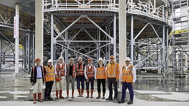 Foto von mehreren Personen, vor einem Gerüst, in Schutzausrüstung. 