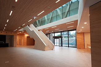 Foto vom inneren eines Gebäudes in der Eingangshalle. In der Mitte erstreckt sich eine Treppe aus Holz.  