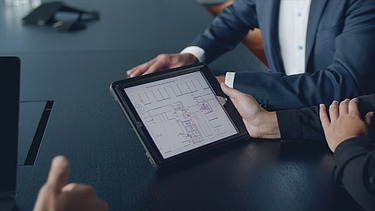 Foto von einem einer Besprechung mit einem Fokus auf ein I-Pad. Auf dem I-Pad ist ein Bauplan zusehen.  