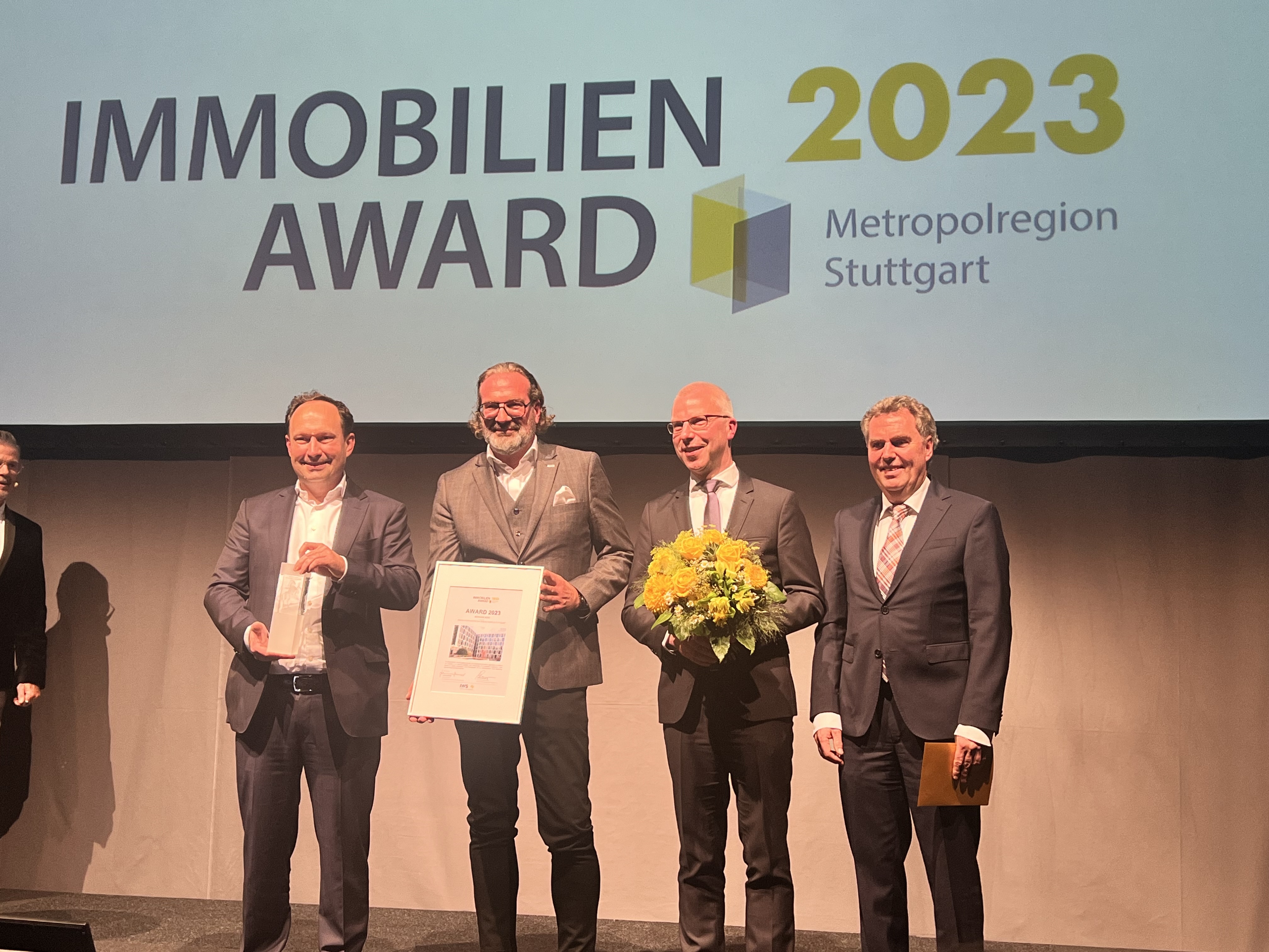 Foto von vier Männern im Anzug die auf einer Bühne stehen. 