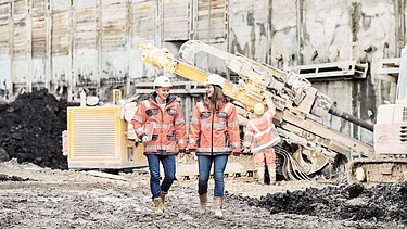 Foto von einem Mann und einer Frau. Beide schauen glücklich. Sie befinden sich auf einer Baustelle. Hinter ihnen steht eine große Maschine.   