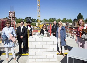 Foto von mehren Personen auf einer Baustelle, sie sind schick gekleidet und haben Kuscheltiere in der Hand. Sie stehen um eine kleine Mauer auf denen ebenfalls Kuscheltiere liegen.  
