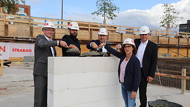 Foto von vier Personen in Anzug, die auf einer kleinen Mauer Beton verteilen. 