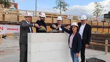 Foto von vier Personen in Anzug, die auf einer kleinen Mauer Beton verteilen. 