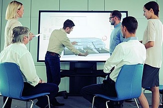 Foto von mehren Personen, die vor einem Fernseher stehen. Es ist eine Präsentation von dem Modele eines Hauses zusehen. Eine Personen verdeutlicht etwas bei der Präsentation.         