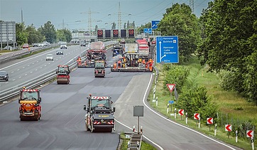 Foto einer Autobahnbaustelle mit mehreren Straßenbaumaschinen darauf, rechts ein blaues Verkehrsschild