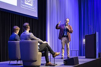 Foto von drei Personen auf einer Bühne, die Person rechts steht, die anderen Beiden sitzen in Couchsesseln