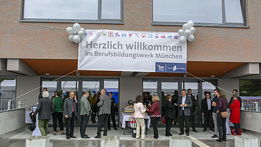 Foto vom Eingang des Berufsbildungswerk München.  Es vorm Eingang. Es herrscht eine ausgelassene Stimmung. 