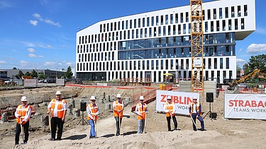 Foto von mehreren Männern in Warnweste auf einer Baustelle. Sie halten einen Spart in der Hand, der in den Boden gesteckt ist. Die Baustelle befindet sich vor einem Bürogebäude. 