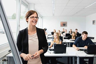 Foto eines Raumes mit mehreren sitzenden Personen, im Vordergrund steht eine weibliche Person mit Brille