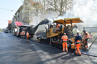 Foto einer Asphalteinbaumaschine im Einsatz, dahinter mehrer Arbeiter