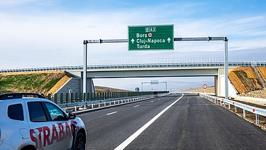 The picture shows a white car with the inscription STRABAG on a motorway, in the background a green traffic sign and a crossing bridge