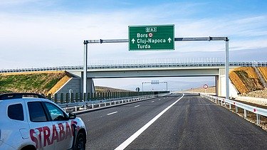 The picture shows a white car with the inscription STRABAG on a motorway, in the background a green traffic sign and a crossing bridge