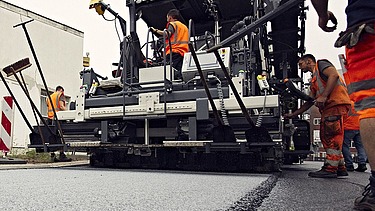 Foto von einer Asphaltmaschine bei der Arbeit, eine Person sitzt in der Maschine, links und rechts stehen noch drei weitere Straßenbauarbeiter