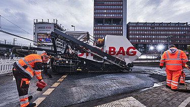 Das Bild zeigt Asphaltierarbeiten mit zwei Arbeitern in Schutzanzügen und einer großen Asphalbaumaschine, im Hintergrund ein Bürogebäude