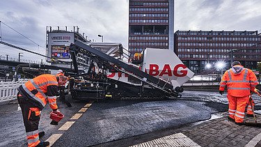 Das Bild zeigt Asphaltierarbeiten mit zwei Arbeitern in Schutzanzügen und einer großen Asphalbaumaschine, im Hintergrund ein Bürogebäude