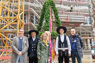 Fünf freundlich schauende Person stehen vor einem Richtkranz  