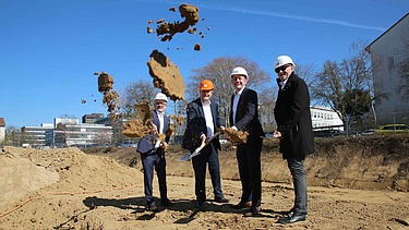 Foto von vier Männern in Anzug und einem Helm auf. Sie sind auf einer Baustelle und schaufeln Erde mit einem Spart.    