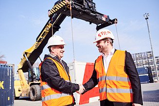 Photo of two people shaking hands and wearing protective clothing, behind which you can see an excavator with its gripper arm extended